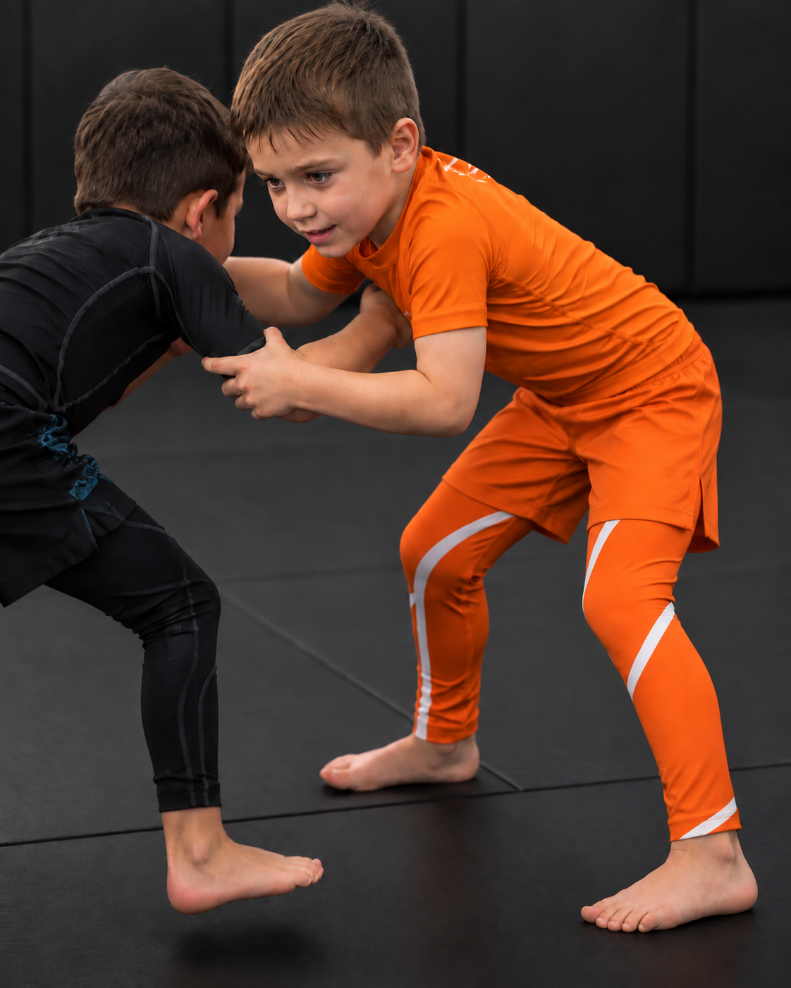 Roll Gear Orange Swirl Spats – Orange/White – Kids, worn during a kids grappling drill on the mat.