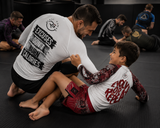 Roll Gear Excuses Don’t Burn Calories Rash Guard – White – Men’s, back view, playing with a child on the mat.
