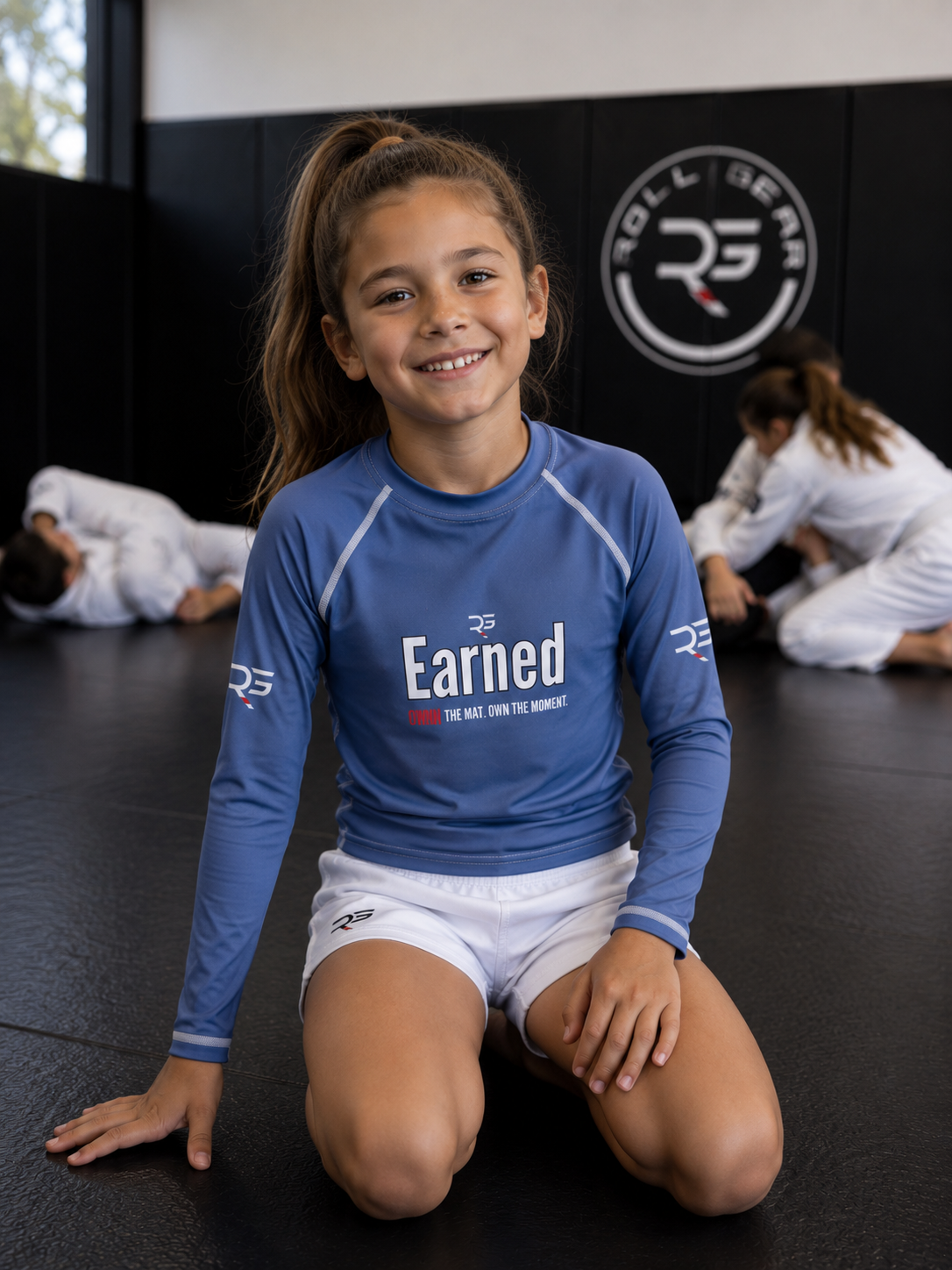 Roll Gear Earned Not Given Long Sleeve Rash Guard – Blue – Kids, smiling girl kneeling on the mat.