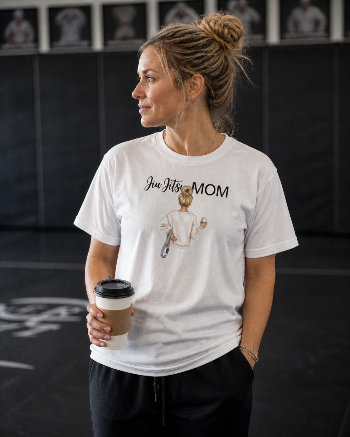 Woman wearing a 'Jiu Jitsu Mom' t-shirt holding a coffee cup in a gym setting.