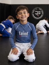 Roll Gear Earned Not Given Long Sleeve Rash Guard – Blue – Kids, smiling boy kneeling on the mat in gi pants.