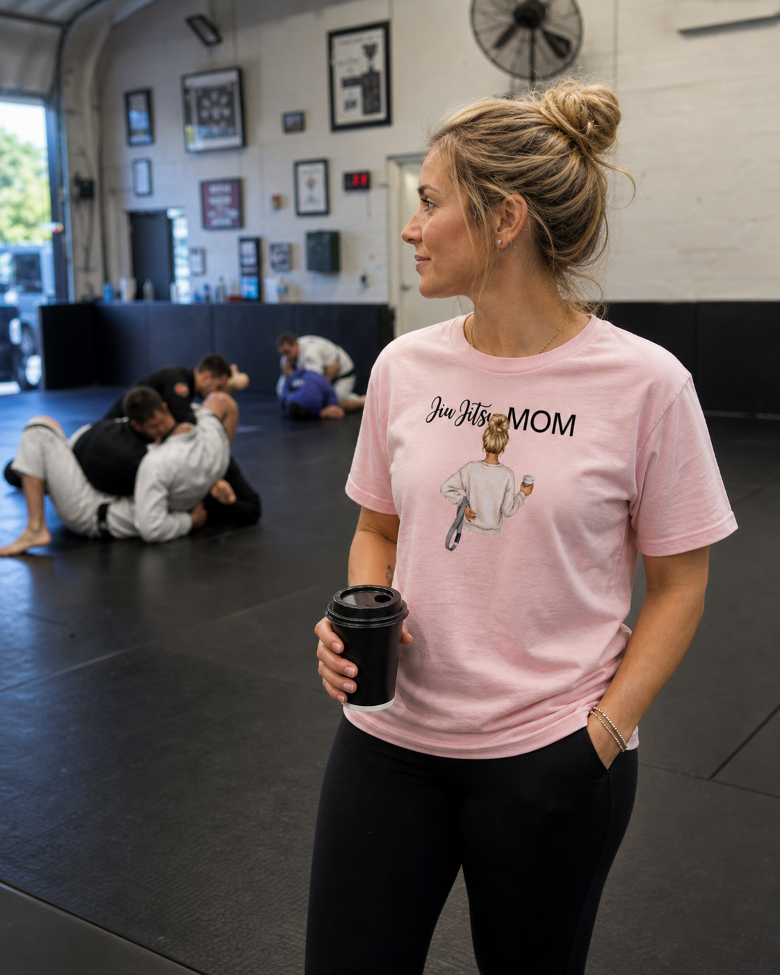 Woman in a gym wearing a pink 'Jiu Jitsu Mom' t-shirt holding a coffee cup.