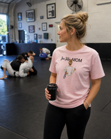 Woman in a gym wearing a pink 'Jiu Jitsu Mom' t-shirt holding a coffee cup.