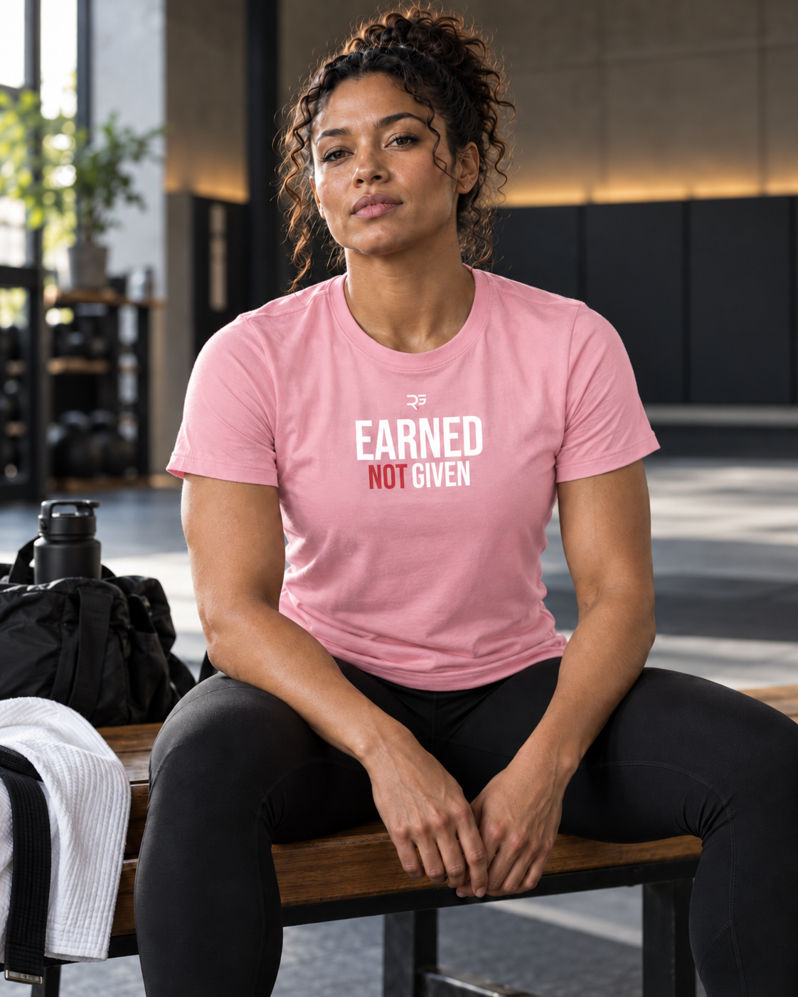 Roll Gear Women's Earned Not Given Motivational T-Shirt, worn seated on a gym bench.