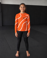 Roll Gear Orange Swirl Long Sleeve Rash Guard – Orange/White – Youth, front view on a girl standing on the mat.