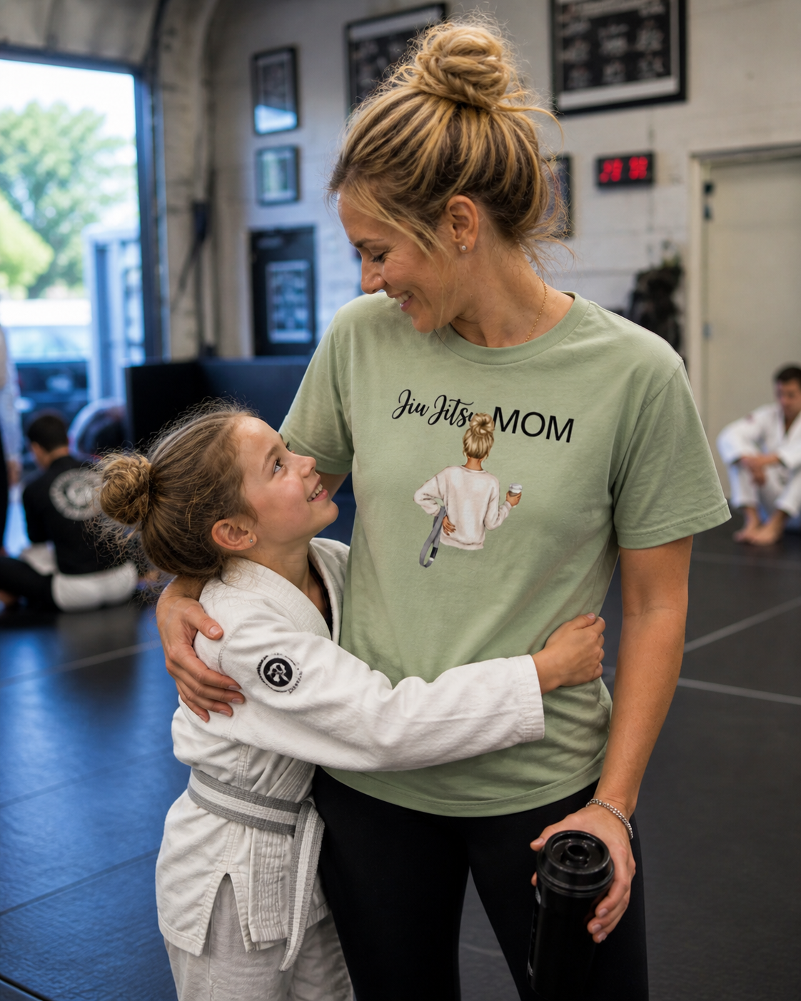 Woman in 'Jiu Jitsu Mom' t-shirt hugging a child in a martial arts uniform in a gym.