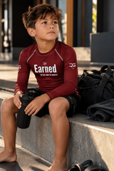 Roll Gear Earned Not Given Long Sleeve Rash Guard – Deep Red – Kids, boy seated by a gym bag.