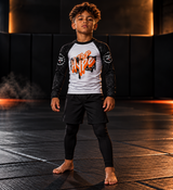 Roll Gear Tap Hype Long Sleeve Rash Guard – Orange & White – Youth, front view on a boy standing on the mat.