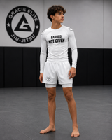 Person wearing a white 'Earned Not Given' shirt and shorts in front of a Gracie Elite Jiu-Jitsu logo.
