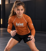 Roll Gear Earned Not Given Long Sleeve Rash Guard – Burnt Orange – Youth, worn by a girl in a grappling stance.