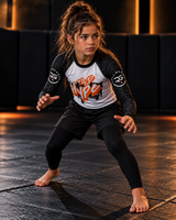 Roll Gear Tap Hype Long Sleeve Rash Guard – Orange & White – Youth, worn by a girl in a grappling stance.