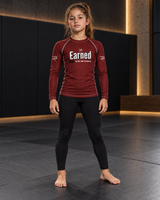 Roll Gear Earned Not Given Long Sleeve Rash Guard – Deep Red – Youth, girl standing on the mat, front view.