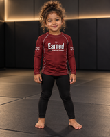 Roll Gear Earned Not Given Long Sleeve Rash Guard – Deep Red – Kids, girl standing on the mat, front view.