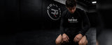 A man in a black Roll Gear long-sleeve compression shirt and shorts kneeling on a dark gym floor, with the Roll Gear logo visible on the wall behind him.