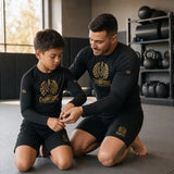 A man and a boy kneeling on a gym floor, both wearing matching black long-sleeve rash guards and shorts with a gold 'Champions' crest logo. The man is adjusting the boy's rash guard in a gym setting with kettlebells and foam rollers visible in the background.