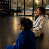Two people in martial arts uniforms sitting on a mat inside a dojo, with large windows showing trees and a vehicle outside.