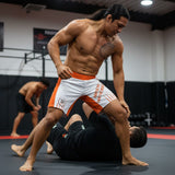 Roll Gear white athletic performance shorts with orange side panels and waistband. Features text "OWN THE MAT. OWN THE MOMENT."