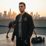 Roll Gear black polyester bomber jacket with embroidered logo, worn by a man on a rooftop at sunset.