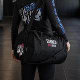A black duffel bag with white and pink text graphics, worn crossbody by a person in a gym setting. The bag features the text 'EARNED NOT GIVEN' on the front.