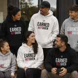 A family of six wearing matching custom hoodies with the text 'EARNED NOT GIVEN' in black and red. The group includes a mother and father, a daughter, and two sons, each wearing a hoodie in black, white, or gray, standing and sitting in front of a modern building.