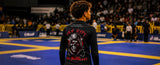 A person standing on a blue and yellow wrestling mat, viewed from behind, wearing a black long-sleeve shirt with a large red and white graphic print on the back reading 'TAP HYPE' and 'BOGXXX'.