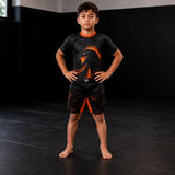 A young boy stands on a black padded martial arts mat wearing a black short-sleeve rash guard with a fiery orange dragon graphic and matching black rash guard shorts with orange side panels.