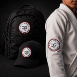 A black backpack and black baseball cap with matching circular 'ROLL GEAR RG' patches are shown beside a person wearing a white gi jacket, all on a dark studio background.