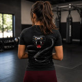 A woman viewed from behind in a gym, wearing a black short-sleeve athletic shirt with a large graphic of a coiled black snake and an open snarling snake head on the back, paired with burgundy leggings.