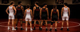 Eight male wrestlers in singlets stand in a line on a wrestling mat, viewed from behind. Four wear black singlets and four wear white singlets, both printed with red text reading 'JUST THE DEED / CAN'T THE SPION'.
