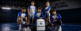 Five athletes in blue and white wrestling uniforms pose with a white and blue wrestling bag on a dark blue wrestling mat inside a gym. A banner in the background reads 'ONE TEAM ONE STANDARD'.