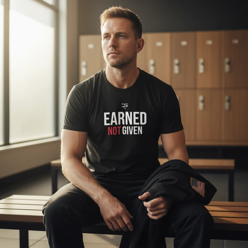 Roll Gear Earned Not Given black heavyweight cotton t-shirt with white and red graphic print on chest. Model sitting in locker room.