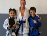 A woman in a white martial arts gi with a blue belt stands between two children wearing white and blue gi uniforms with black belts. The child on the left holds a black plaque, and the child on the right gives a thumbs-up.