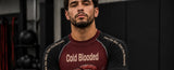 A man wearing a Cold Blooded brand athletic long-sleeve shirt in a gym setting. The shirt features a dark maroon chest and black sleeves with white text reading 'Cold Blooded' and 'Own the Mind.