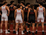 A group of male wrestlers standing in a huddle on a wrestling mat, viewed from behind. They are wearing singlets in white and black with red trim and the text 'OWN THE MAT.