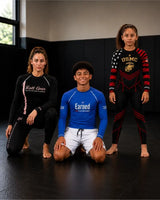 A woman in a white martial arts gi with a blue belt stands between two children wearing white and blue gi uniforms with black belts. The child on the left holds a black plaque, and the child on the right gives a thumbs-up.