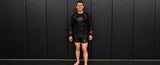 A man wearing a black long-sleeve rash guard and black shorts with red wrist guards, standing against a dark paneled wall.