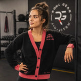 A woman in a gym wearing a black cardigan with pink trim and matching pink cuffs, featuring a small embroidered logo on the left chest. The background shows a dark gym interior with a large 'ROBL GEAR JIU JITSU' logo on a black wall.