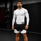 A muscular man in a gym wearing a white long-sleeve compression shirt and black athletic shorts with white panels and a patterned waistband.