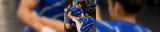 A man in a blue athletic jersey and shorts sitting on a gym floor talking to another person, with a blurred close-up of the blue jersey in the foreground.