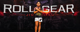 A woman stands in front of a dark promotional backdrop with large white text reading 'ROLL GEAR' and smaller 'JIU JITSU' text. She is wearing a dark patterned short-sleeve top and white athletic shorts with black graphics.