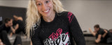 A smiling blonde woman wearing a black long-sleeve athletic shirt with pink and white graphic text, photographed in a gym setting with other people blurred in the background.