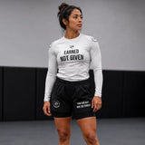 A woman wearing a white long-sleeve athletic shirt with the text 'EARNED NOT GIVEN' and black athletic shorts, standing in a studio setting.
