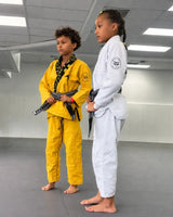 Two youth athletes in BJJ gis with Roll Gear patches, one in yellow and black and one in white with black and yellow trim, standing together on gym mats