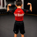 A child viewed from behind wearing a red and black rash guard with 'Cold Blooded' printed across the back, paired with black athletic shorts in a gym setting.