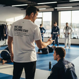 Man in a white Jiu Jitsu Dad tee seen from behind at a gym, showing the Mat Side Coaching Staff back print.
