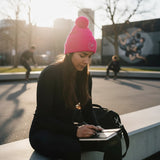 Roll Gear bright pink knit beanie with two pom-poms and embroidered logo, worn by a woman outdoors.
