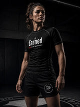 A woman in a dark gym setting wearing a black short-sleeve athletic t-shirt with 'Earned' printed in white on the chest and black athletic shorts with a white circular logo.