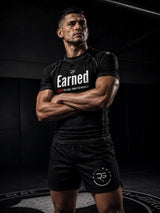 A muscular man in a black athletic gym setting, wearing a black short-sleeve t-shirt with 'Earned' printed in white across the chest and black shorts with a circular logo on the left leg.