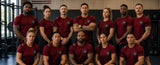 A group of ten fitness athletes posing together in a gym, wearing matching dark red athletic t-shirts with a circular logo on the chest.